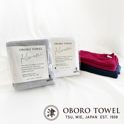兩件獨立包裝的OBORO TOWEL毛巾禮盒，一件為灰色，另一件為白色，均展示其精緻的日式工藝與柔軟觸感。右側堆疊著三條不同顏色的毛巾，分別為深紅色、深藍色和另一條深藍色，質地蓬鬆。