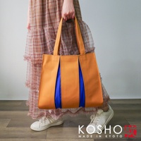 橘色棉帆布扇形托特包，搭配藍色內裡，由手提帶提起，展示於穿著格子洋裝的模特兒身上。