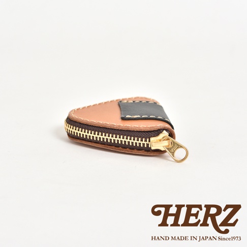 這是一款由日本品牌HERZ職人手作的皮革零錢包，呈現獨特的三角飯糰造型，採用棕色與黑色拼接皮革製成，表面有精緻的手縫線細節，並配有金色的拉鍊設計，右下角帶有HERZ品牌商標與日製字樣。