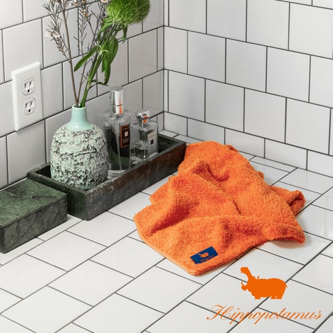 橘色竹棉混紡長毛巾，質地蓬鬆柔軟，帶有深藍色品牌標籤，置於白色磁磚檯面上。