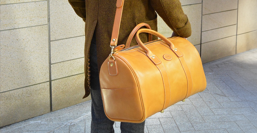 棕色皮革波士頓包，配有可拆卸肩帶和手提把，包身有拉鍊開合，外部設有側面口袋，細節處有縫線裝飾。