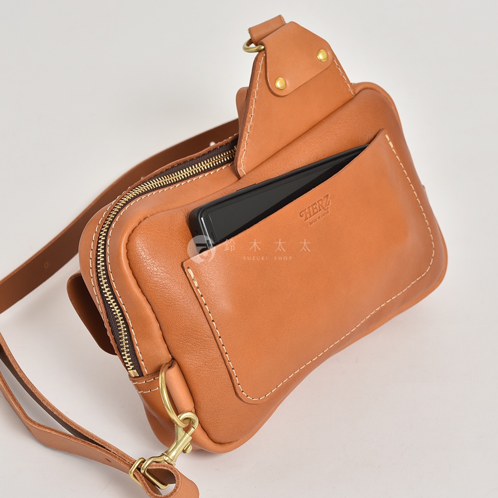 棕色皮革雙口袋斜背包，一個口袋裝有黑色手機，另一個口袋為拉鍊式設計，搭配金屬扣環和可調式肩帶。