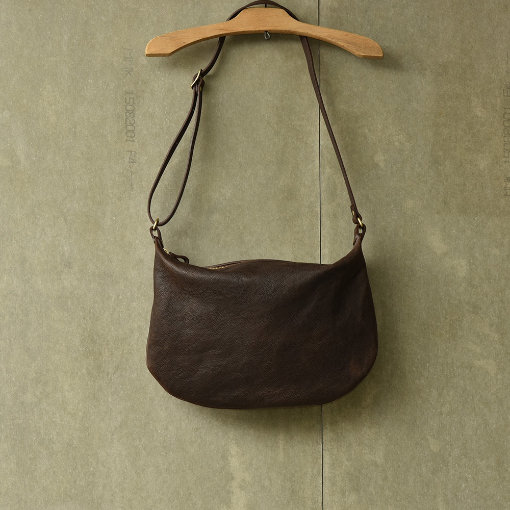 深棕色皮革製成的半月形單肩包，由職人手工製作，附有可調式肩帶，懸掛於木質衣架上。