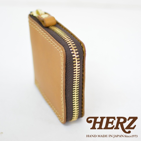側面拍攝的HERZ日本職人手作淺棕色皮革短夾，展示其細膩的縫線工藝與金色拉鍊細節，右下方印有品牌名稱與手工製作標誌。