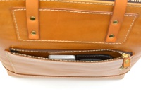 職人手作皮革威靈頓直式拉鍊托特包，採用光滑的棕褐色皮革製成，設有一個橫跨包身前方的拉鍊口袋，口袋內可見一部銀色手機和一個黑色錢包。包包有清晰的縫線細節和金屬釘。