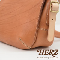 職人手作皮革郵差包，採用自然色澤的牛革，搭配古銅色金屬扣環與淺色系皮革背帶，車縫線清晰，展現皮革天然紋理。