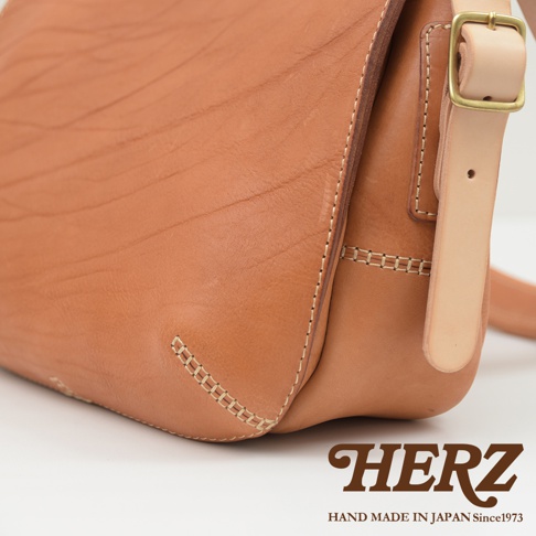 職人手作皮革郵差包，採用自然色澤的牛革，搭配古銅色金屬扣環與淺色系皮革背帶，車縫線清晰，展現皮革天然紋理。