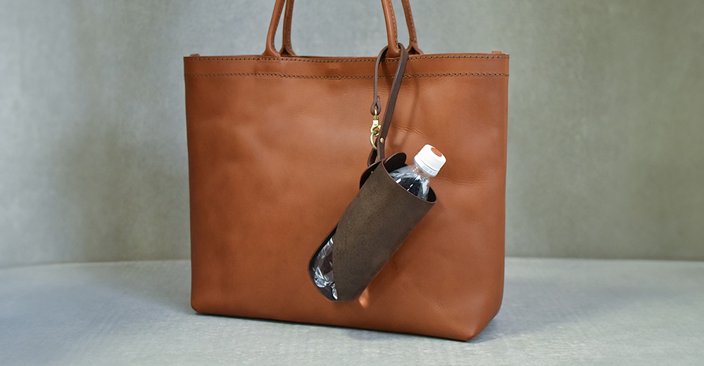 職人手作皮革Organ水壺套，深棕色皮革，附有掛繩及金屬扣環，可掛於棕色皮革托特包上，內裝有白色瓶蓋的寶特瓶。