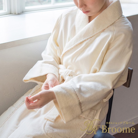 一位女性穿著米白色今治純棉浴袍，繫著腰帶，坐在椅子上，浴袍領口和袖口有細緻的滾邊裝飾，材質看起來柔軟舒適。