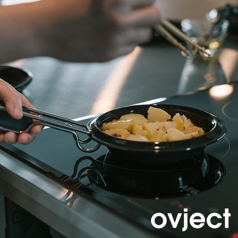 一個黑色的 25 公分琺瑯煎鍋，鍋內裝有切塊的馬鈴薯，正被放在電磁爐上加熱。鍋柄為金屬材質，配有黑色防滑握把。