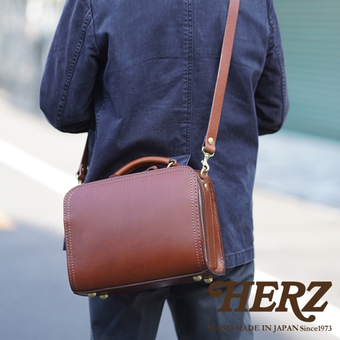 職人手作皮革兩用方形單肩包，採用棕色皮革製成，帶有手提把及可拆卸肩帶，金屬拉鍊與底部金屬釘飾，整體風格簡約。