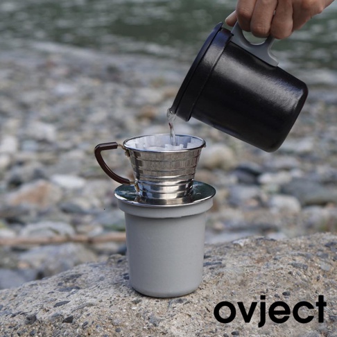 一個銀色金屬濾杯，帶有棕色把手，置於灰色杯子上。一個黑色水壺正將熱水倒入濾杯，濾杯內有咖啡濾紙。