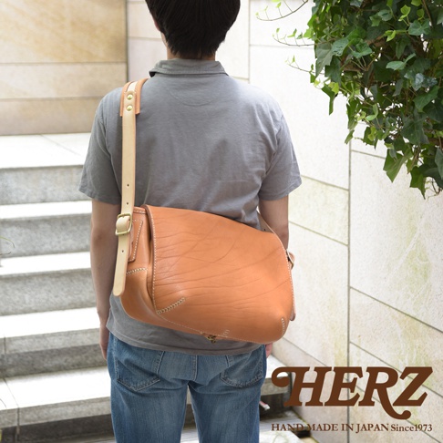 職人手作皮革郵差包，淺棕色，帶有天然皮革紋理，以金屬扣環開合，寬版皮革背帶可調整長度，包身縫線明顯，整體呈現復古風格。