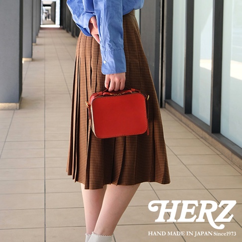 日本HERZ 職人手作皮革兩用迷你小方包，展示紅色皮革包款，搭配棕色格紋裙，強調日本製造since1973