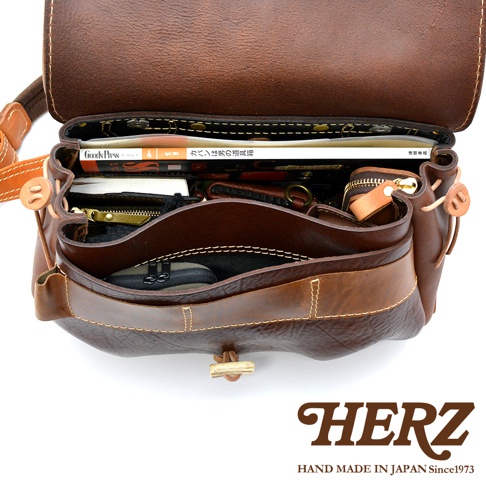 職人手作的棕色皮革郵差包，內部裝有雜誌、文件夾、錢包和化妝包，包身有鹿角造型的扣繩設計。