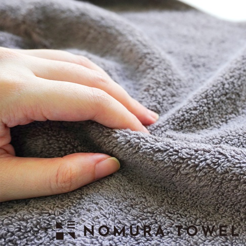 野村毛巾品牌標誌和「NOMURA TOWEL」文字，以特寫鏡頭展示深灰色毛巾柔軟的紋理。