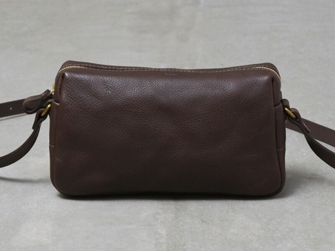 深棕色皮革製成的職人手作輕巧拉鍊單肩包，包身呈現圓潤的長方形狀，配有金色拉鍊和皮革肩帶。