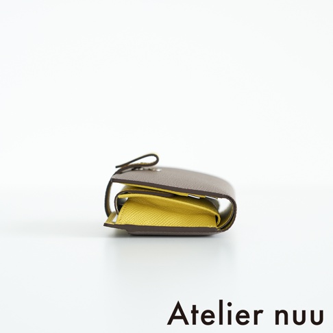 Atelier nuu 品牌下，一張灰褐色與鵝黃色皮革對折短夾的側邊拍攝圖，短夾下方右側有 Atelier nuu 的品牌文字。