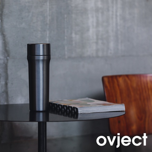 黑色金屬三用保溫瓶，瓶身光滑，帶有銀色瓶蓋，置於黑色圓形桌上，旁邊有一本彩色封面雜誌和木質椅子。