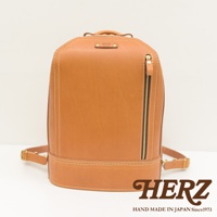 一個淺棕色皮革製的立體商務後背包，有著雙肩帶和一個垂直的拉鍊口袋，整體設計簡潔，展現職人手作質感。