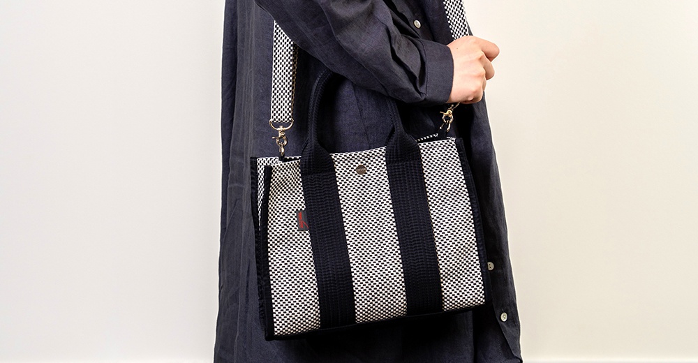 一款藍白條紋的刺子織方形托特包，配有黑色織帶提把與可拆卸斜背帶，包身一側有紅色標籤。