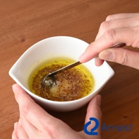 一隻手拿著不鏽鋼萬用醬料小勺，正在白色碗中攪拌油醋醬，醬料中含有黃色油體和褐色芥末籽。