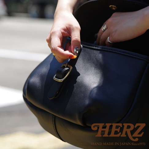 日本HERZ手工皮革包，特寫女性手指正在打開包包的搭扣，黑色皮革材質，旁邊有品牌標誌。