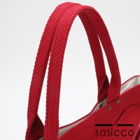 紅色三河木綿刺子織托特包，特寫兩條厚實的紅色提把，提把有明顯的織紋。包體邊緣可見金屬鉚釘細節。