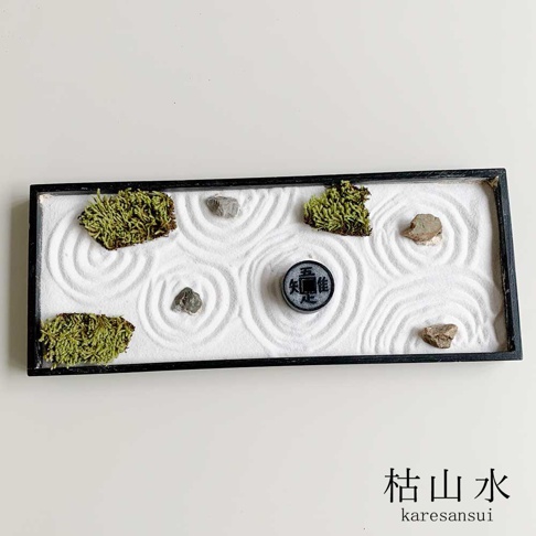 黑框長方形枯山水模型，內有白色細沙，以耙子畫出漣漪紋路，四角和中間各置一塊灰色小石塊，其中央處有一枚有文字的圓形裝飾物，模型兩側各有一簇深綠色苔蘚。