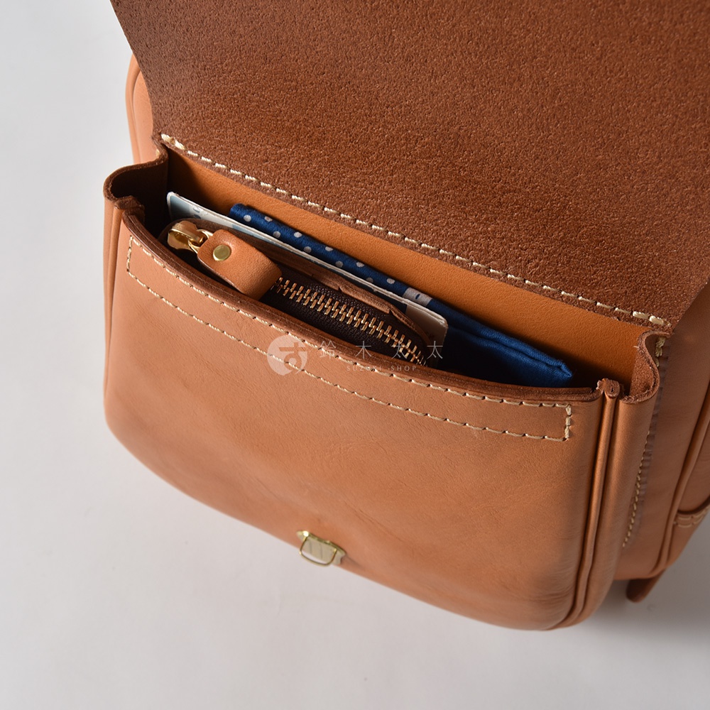 淺棕色皮革迷你後背包，翻蓋設計，前方立體口袋內有拉鍊小包、藍色碎花布料和卡片。