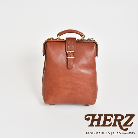 一款棕色皮革迷你軟皮杜勒斯包，帶有金色搭扣和提把，產品標誌為HERZ，Hand Made in Japan Since 1973。