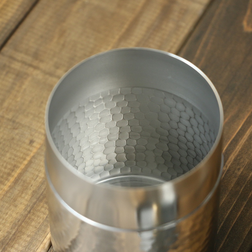 圓柱形不鏽鋼茶葉罐，表面有錘目紋凹凸紋理，內壁亦有相同的蜂窩狀壓紋，呈現銀灰色金屬光澤，置於木紋背景上。