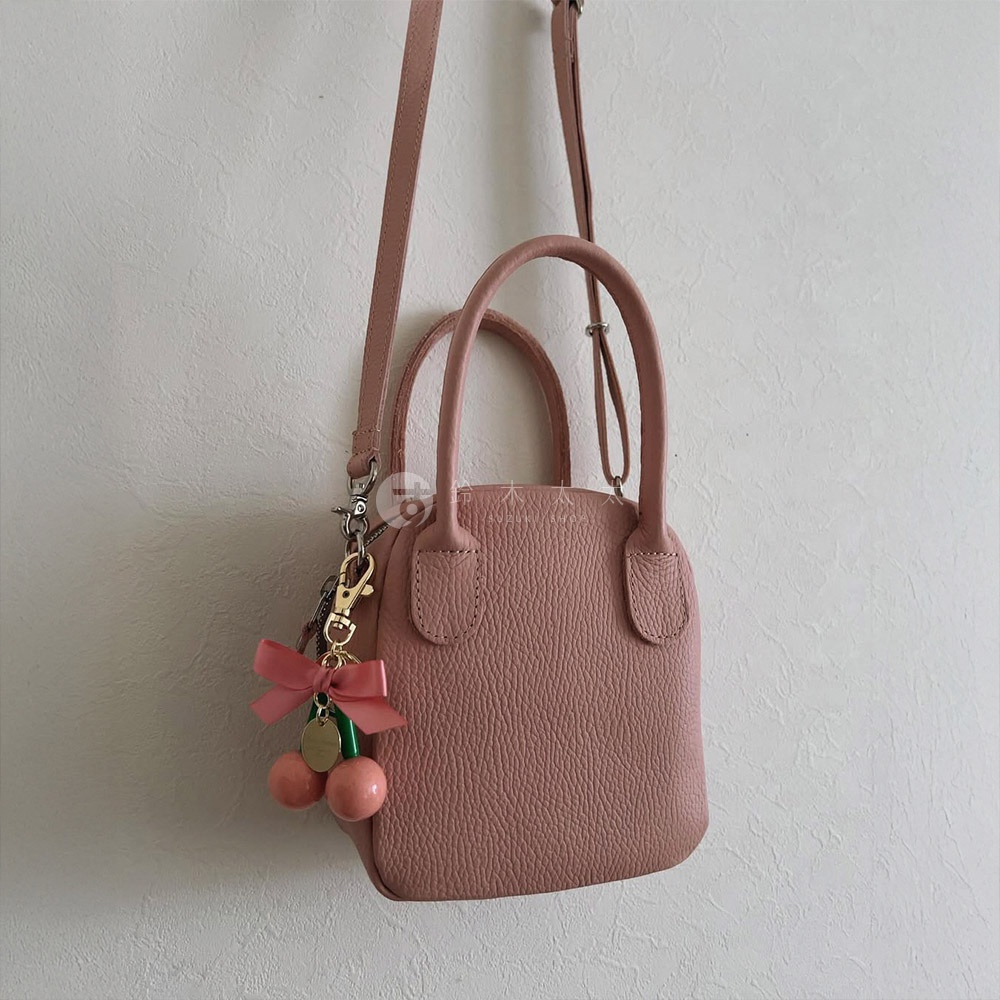 粉紅色荔枝紋皮革手提包，可斜背，包包上有兩條手提帶，左側掛有粉紅色蝴蝶結與兩顆粉紅色櫻桃吊飾。