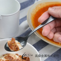 一只手正拿著一把日製熔岩黑石水果甜點匙，匙中盛有優格和麥片。背景為白色餐具和橘色食物。