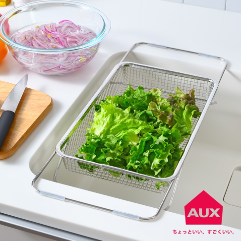 不鏽鋼伸縮瀝水籃內盛裝著新鮮的綠葉生菜，籃架置於水槽內，旁邊有裝有紫洋蔥的玻璃碗和一把刀。