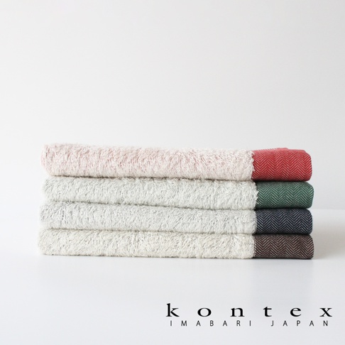 四條堆疊的毛巾，材質為長纖有機棉，呈現斜紋編織，分別為淡粉色、淺灰色、深灰色和淺棕色，每條毛巾的一端帶有不同顏色的織邊。