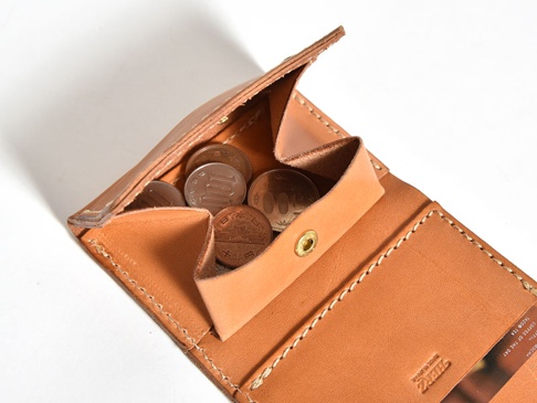 職人手作皮革三折式迷你短夾，採用原色植鞣皮革製成，內含日圓硬幣，錢夾有明顯的縫線細節，並壓印有品牌標誌。