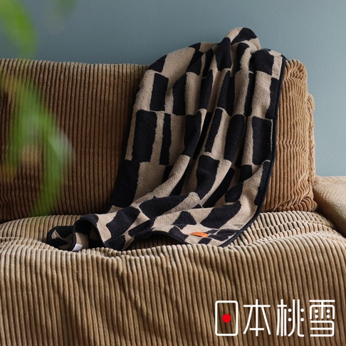 一件鋪在褐色燈芯絨沙發上的格紋浴巾，上面有黑色和米色的幾何圖案，浴巾右下角有「日本桃雪」的紅色標誌。