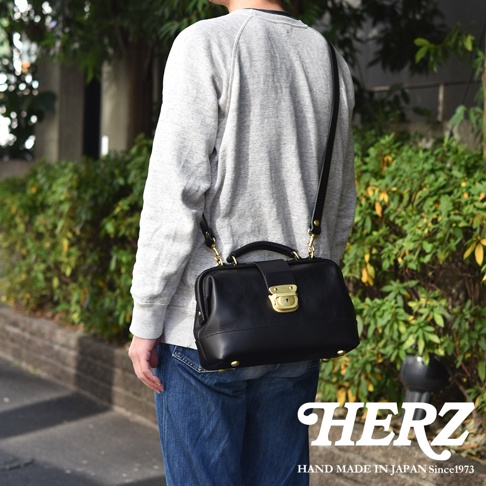 一位穿著灰色長袖上衣與牛仔褲的男子，正背著一款由日本職人品牌 HERZ 手作的黑色皮革迷你口金側背包，包身搭配金色金屬鎖扣，呈現復古簡約質感。