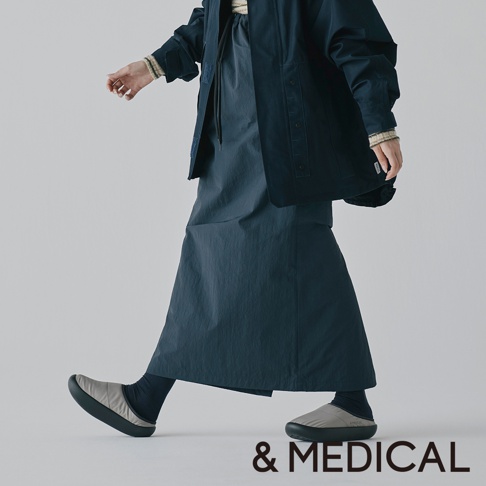 &MEDICAL 的 KAMOLEG 3.0 美腳健身機能鞋 M，展示一位穿著黑色外套、深藍色長裙和灰色舒適拖鞋的人。