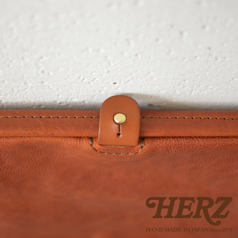 職人手作皮革Organ手拿包G-38，特寫皮革材質的翻蓋扣，附有金屬按鈕，皮革顏色為棕褐色，縫線精緻。