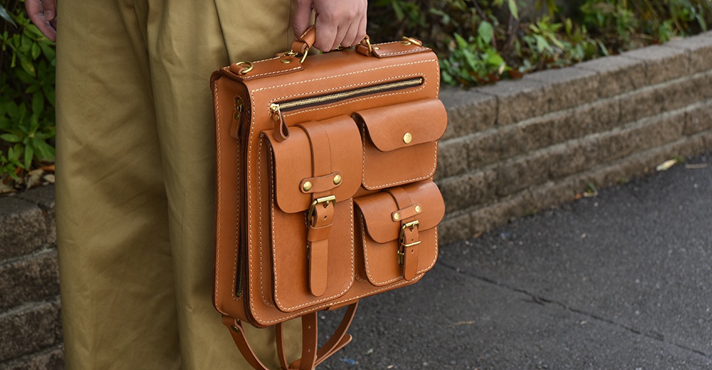 職人手作皮革三口袋三用後背包，採用棕褐色皮革，設有頂部提把、拉鍊主袋、三個前置口袋，其中兩個有磁扣，另一個有皮帶與銅釦設計，並有側面拉鍊及可見的肩帶。