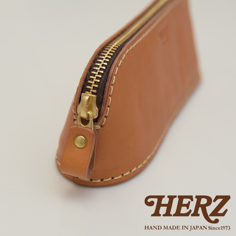 職人手作咖啡色皮革三角弧形收納筆袋，配有黃銅色拉鍊與金屬鈕扣，車縫線清晰可見。