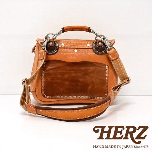 職人手作皮革鹿角扣繩郵差包，主體為淺棕色皮革，中央拼接深棕色皮革，搭配銀色金屬扣環與可調整的皮革背帶，風格簡約。