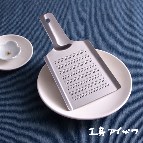 一個銀色的不鏽鋼磨泥器，設有帶有許多小孔的磨板和一個長柄，放在一個白色盤子上，盤子放在藍色織物背景上。
