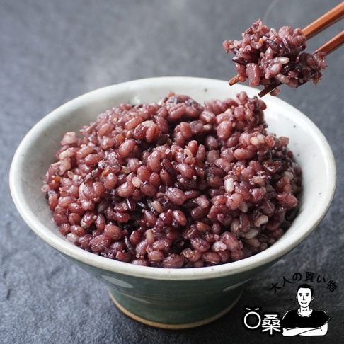 一碗盛滿的深紅色日本國產十六穀米，由木質筷子夾起一撮，米飯顆粒飽滿，呈現多種穀物的混合色澤。