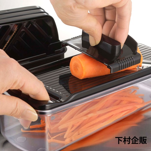 黑色塑膠製四合一刨絲器，正在刨一根橙色胡蘿蔔，刨出的胡蘿蔔絲堆積在透明塑膠集料盒中。