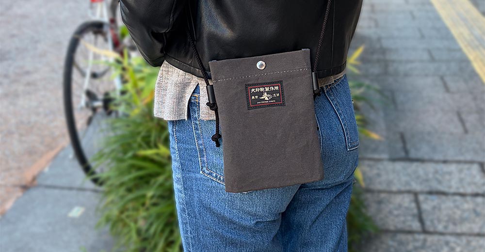 模式B： 一位女性穿著黑色皮衣和牛仔褲，側身背著一個深灰色的帆布手機包，包上有「犬印鞄製作所」的日文標籤，背景是模糊的街道和自行車。
