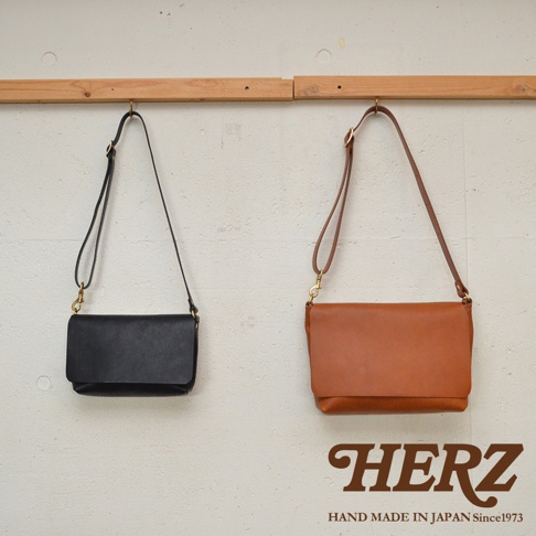 黑色和棕褐色皮革製的兩款HERZ斜背包，皆掛於木製掛鉤上。