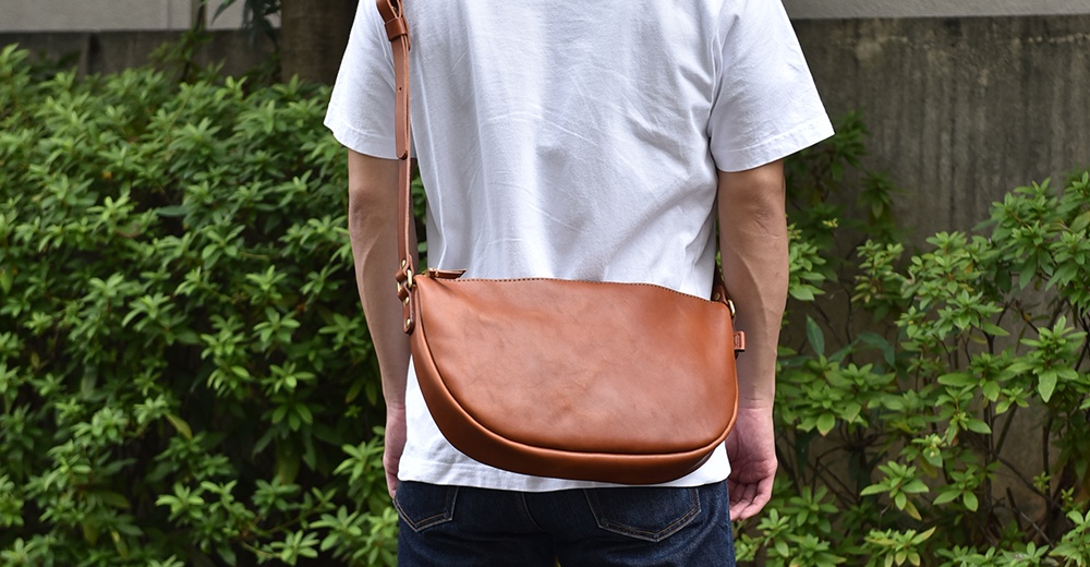 日本HERZ職人手作皮革半圓馬鞍包，展示了棕色皮革材質的光澤與質感，包款的弧形設計與斜背的穿戴方式。